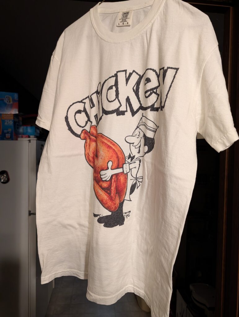 A white t-shirt with a black and white cartoon chef holding a colored realistic looking cooked chicken.